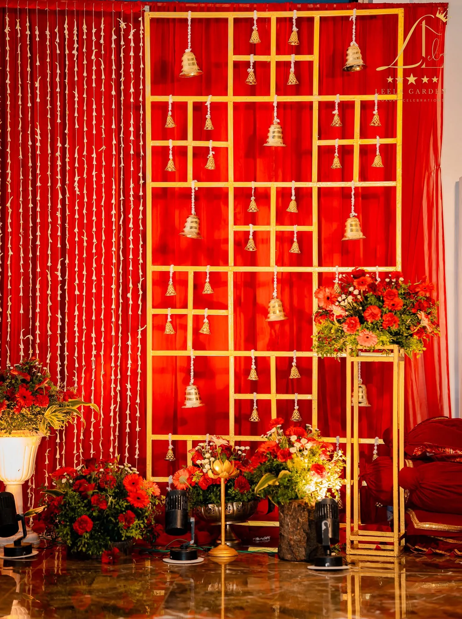 A red and gold ceremonial backdrop hung with brass bells and marigold, framed by floral urns.