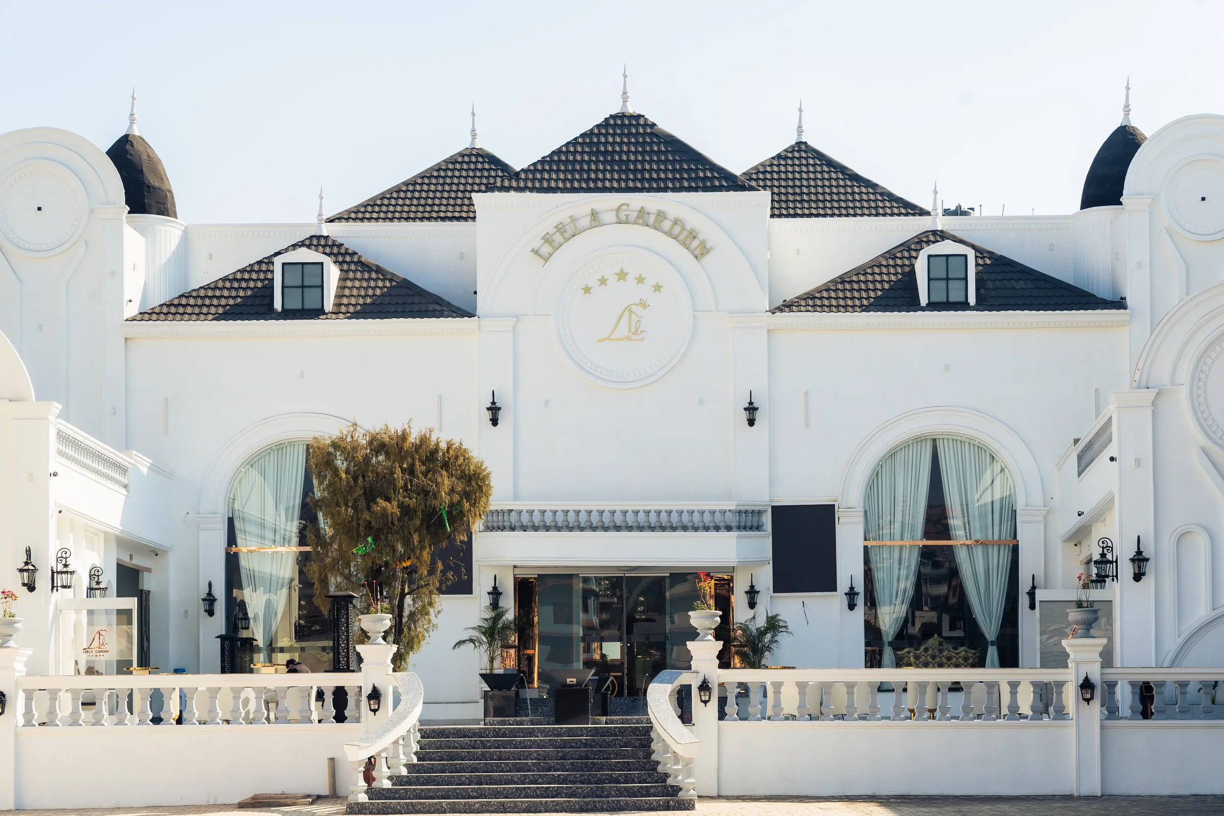 The white palatial facade of Leela Garden in daylight, with its crested Leela Garden signage, arched windows and balustraded entrance stair.