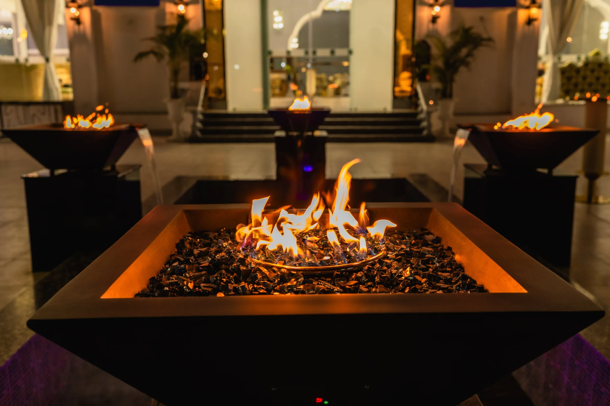 Open fire bowls burning on the outdoor terrace at night, with palms and the lit facade behind them.