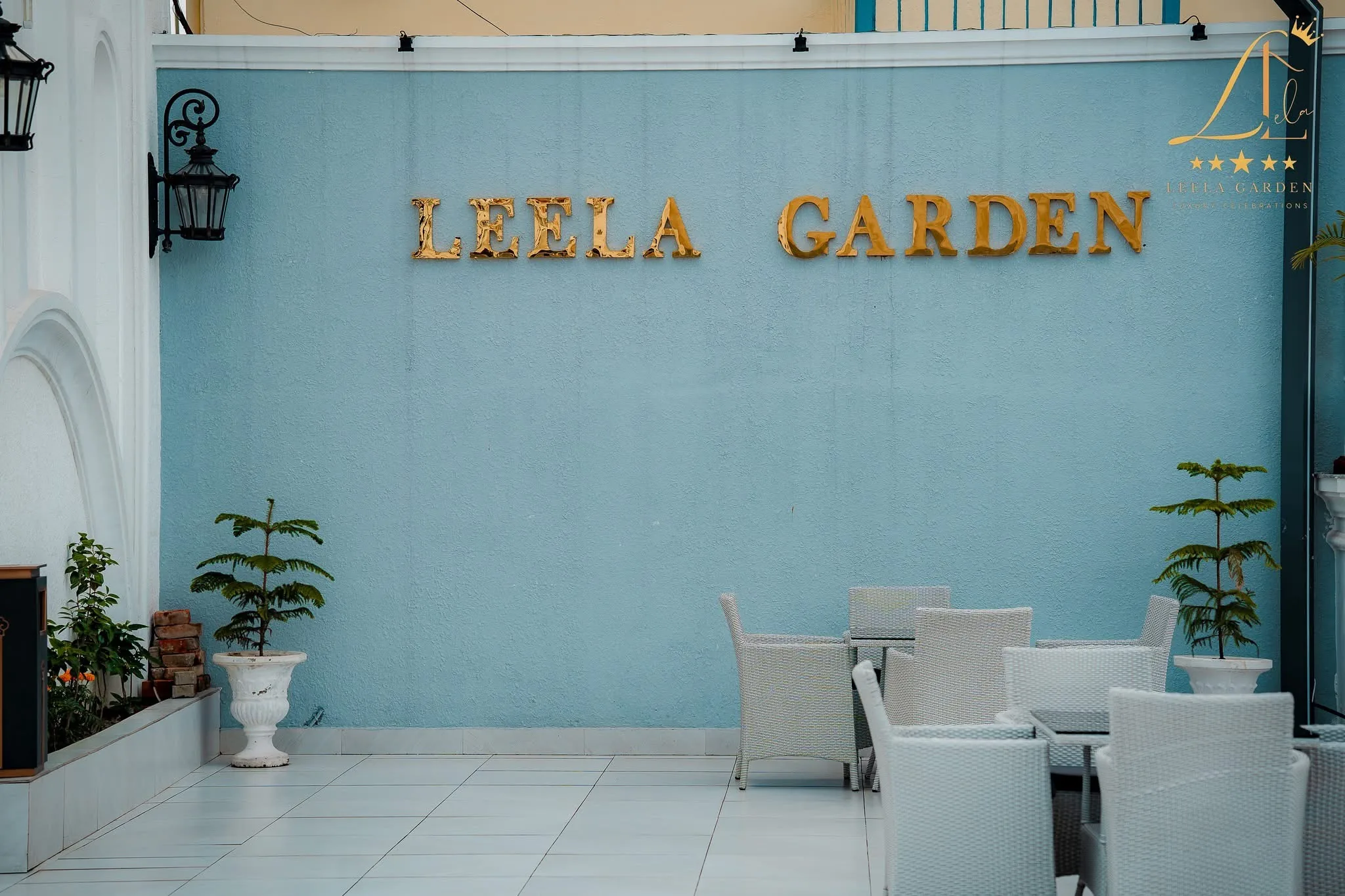 Gold LEELA GARDEN lettering mounted on a pale blue exterior wall above woven lounge chairs.
