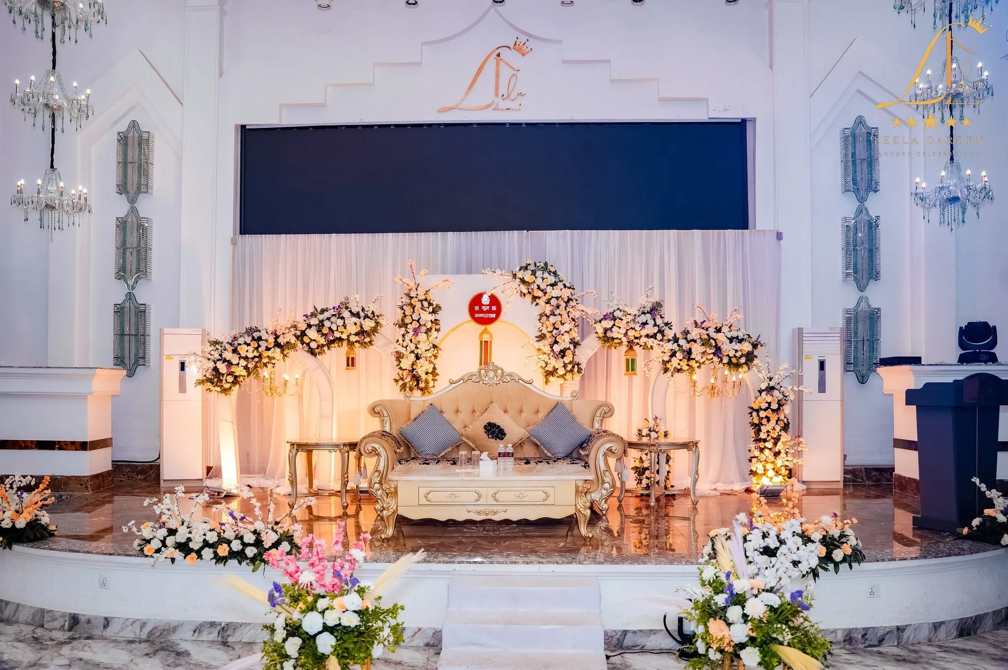 A wedding stage framed by a white and gold floral arch around a gilded settee, the LED wall lit blue behind.