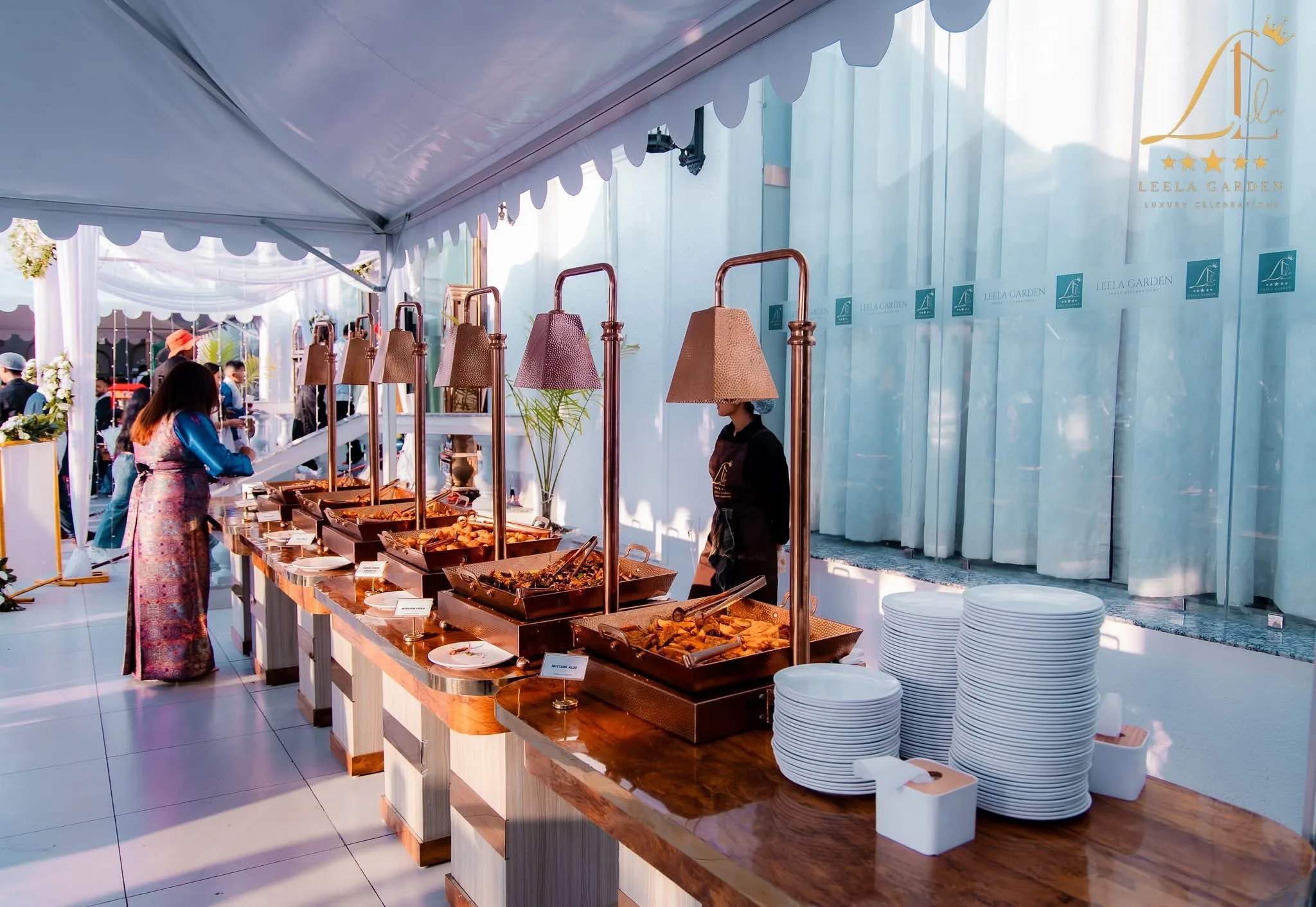 A buffet line set under a white marquee, brass lamps above stacked plates.