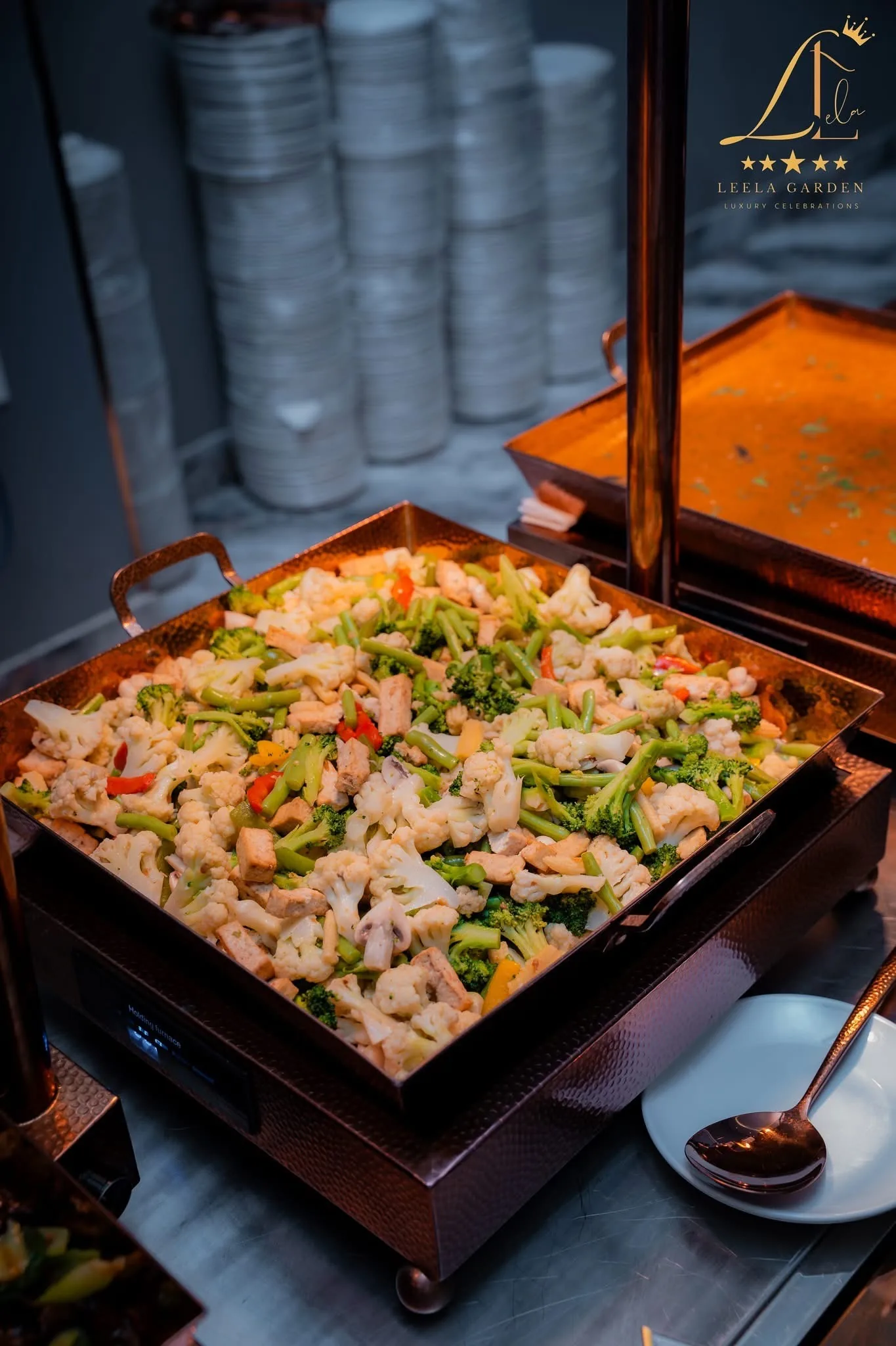 A copper tray of mixed vegetables, stacked plates behind it.
