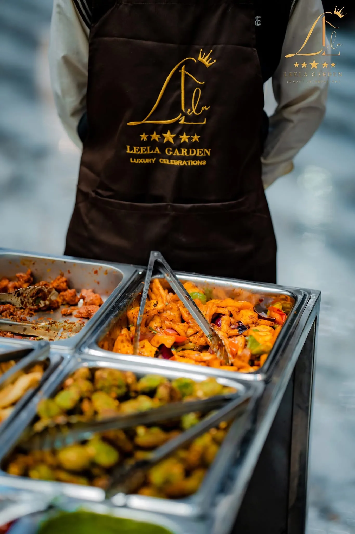 A chef in a Leela Garden apron standing over chafing trays of freshly served food.