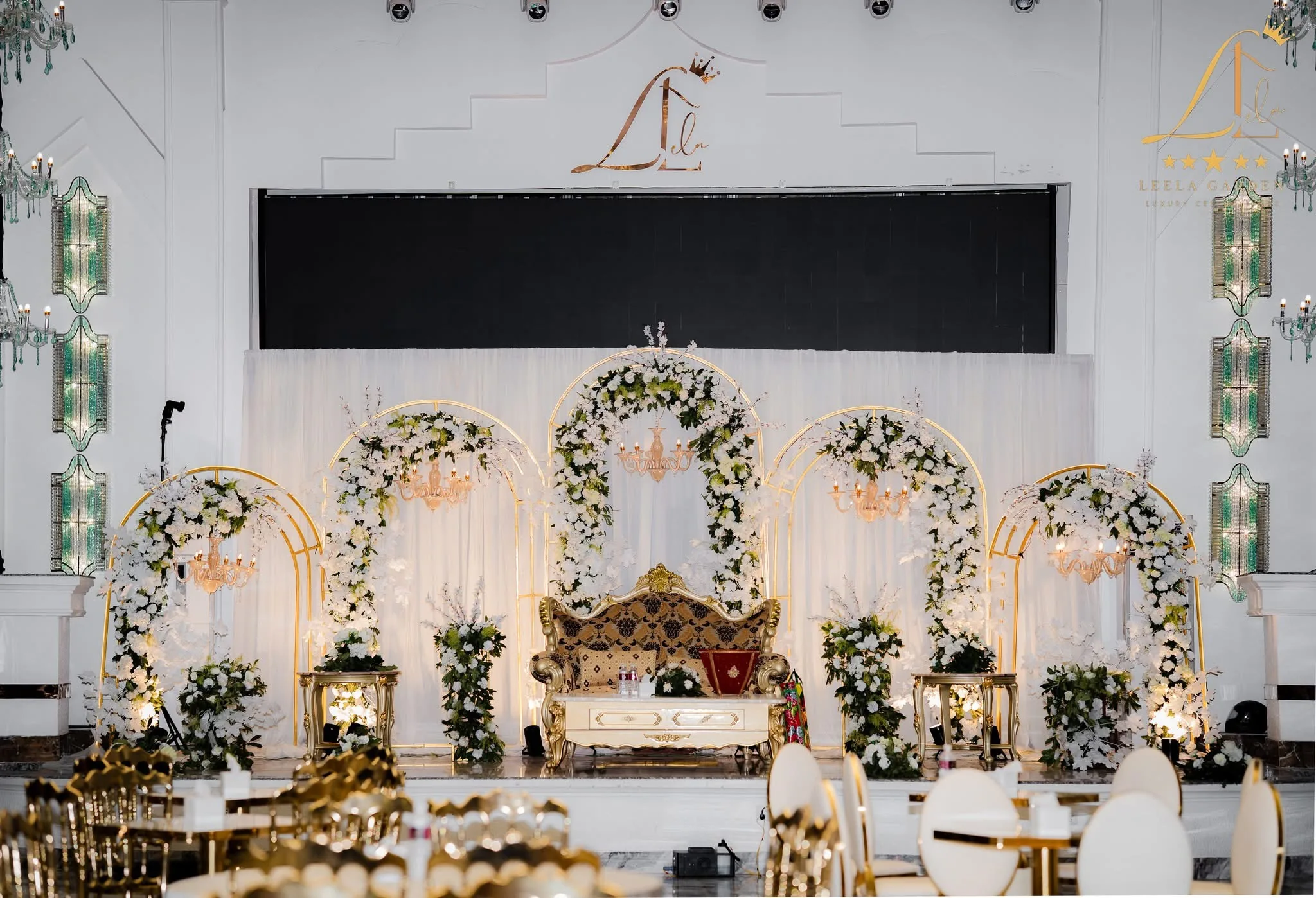 A wedding stage at Leela Garden dressed with white and green floral arches around a gilded ceremonial settee, backed by the hall’s wide LED screen.